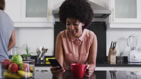 Mujer-Afroamericana-Sosteniendo-Una-Taza-De-Café-Usando-Un-Teléfono-Inteligente-En-La-Cocina-De-Casa