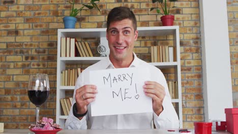 Caucasian-man-making-video-call-holding-handwritten-sign-making-marriage-proposal-and-celebrating-ac