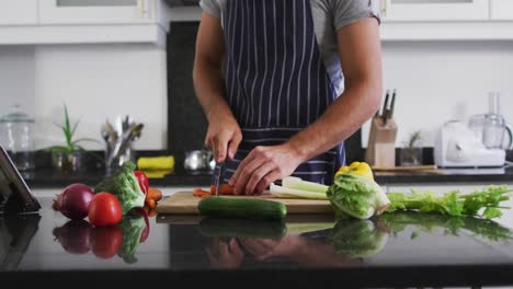Mid-section-of-caucasian-man-wearing-apron-chopping-vegetables-in-the-kitchen-at-home