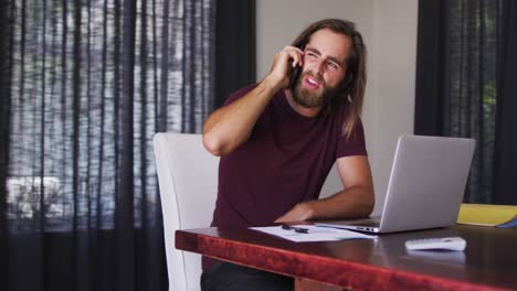 Caucasian-man-talking-on-smartphone-while-using-laptop-at-home