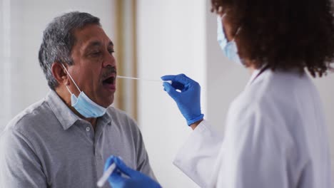 Mixed-race-female-doctor-wearing-mask-doing-swab-test-on-senior-man-at-home