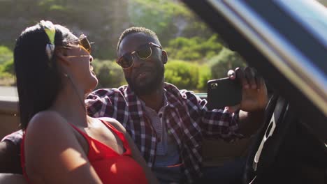 Afroamerikanisches-Paar-Benutzt-Smartphone,-Während-Es-Im-Cabrio-Auf-Der-Straße-Sitzt