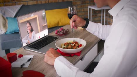 Diverse-couple-on-a-valentines-date-video-call-woman-on-laptop-screen-smiling-man-eating-meal