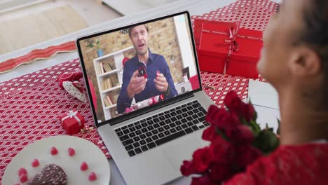 Diverse-couple-on-a-valentines-date-video-call-man-on-screen-showing-ring-to-woman-holding-flowers