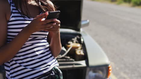 Eine-Afroamerikanische-Frau-Nutzt-Ihr-Smartphone,-Während-Sie-Auf-Der-Straße-Neben-Ihrem-Kaputten-Auto-Steht