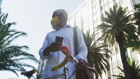African-american-senior-man-wearing-face-mask-with-bicycle-using-smartphone-and-looking-around-in-co