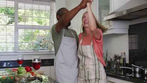 Vielfältiges-älteres-Paar-Mit-Schürzen,-Das-Beim-Zubereiten-Von-Speisen-In-Der-Küche-Tanzt