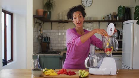 Portrait-of-mixed-race-woman-putting-chopped-fruits-in-juice-maker-in-the-kitchen-at-home