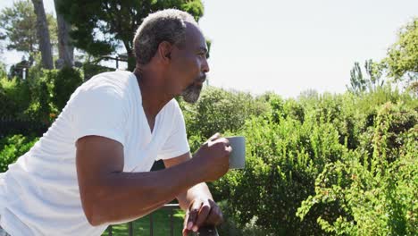 Älterer-Afroamerikanischer-Mann,-Der-An-Einem-Sonnigen-Tag-Auf-Dem-Balkon-Steht-Und-Kaffee-Trinkt