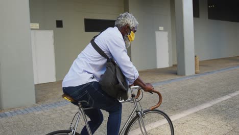 Afroamerikanischer-älterer-Mann-Mit-Gesichtsmaske,-Der-Im-Firmenpark-Fahrrad-Fährt
