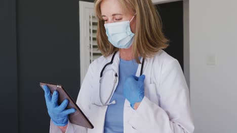 Caucasian-senior-female-doctor-wearing-surgical-gloves-and-face-mask-using-tablet-computer