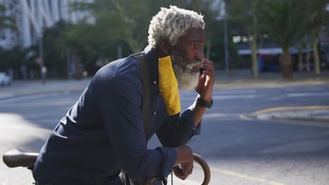 Afroamerikanischer-älterer-Mann-Mit-Gesichtsmaske,-Der-Auf-Dem-Smartphone-Spricht,-Während-Er-Sich-Auf-Sein-Fahrrad-Stützt