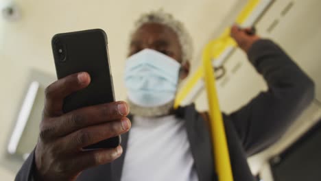 Afroamerikanischer-älterer-Mann-Mit-Gesichtsmaske-Und-Smartphone,-Während-Er-Im-Bus-Steht