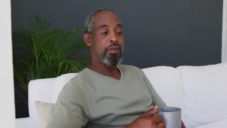 Älterer-Afroamerikanischer-Mann-Hält-Kaffeetasse,-Während-Er-Zu-Hause-Auf-Der-Couch-Sitzt