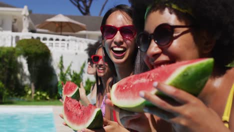 Diverse-group-of-female-friends-eating-watermelon-sitting-at-the-poolside-talking