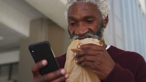 Afroamerikanischer-älterer-Mann,-Der-Im-Firmenpark-Einen-Snack-Isst-Und-Sein-Smartphone-Benutzt