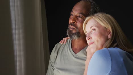 Mixed-race-senior-couple-embracing-each-other-while-looking-out-of-the-window-at-home