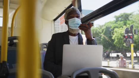 Afroamerikanischer-älterer-Mann-Mit-Gesichtsmaske,-Der-Im-Sitzen-Auf-Dem-Smartphone-Spricht-Und-Einen-Laptop-Benutzt