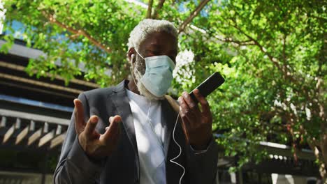 African-american-senior-man-wearing-face-mask-talking-on-smartphone-in-corporate-park