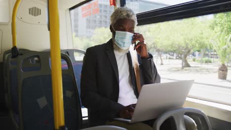 African-american-senior-man-wearing-face-mask-talking-on-smartphone-and-using-laptop-while-sitting-i
