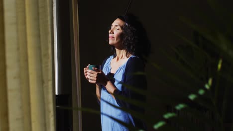 Mixed-race-woman-holding-coffee-cup-while-standing-near-the-window-at-home