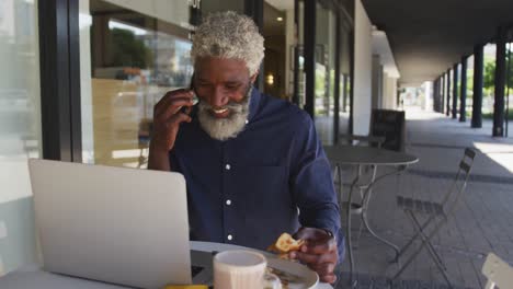 Afroamerikanischer-älterer-Mann,-Der-Auf-Dem-Smartphone-Spricht,-Während-Er-Draußen-Im-Café-Frühstückt