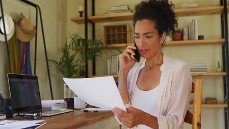Mixed-race-woman-holding-a-document-talking-on-smartphone-while-working-from-home