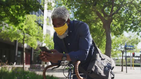 Afroamerikanischer-älterer-Mann-Mit-Gesichtsmaske-Und-Smartphone,-Während-Er-Sich-Auf-Seinem-Fahrrad-Auf-Der-Rechten-Seite-Lehnt