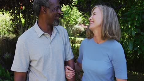 Diverse-senior-couple-walking-in-their-garden-arm-in-arm-in-the-sun-smiling