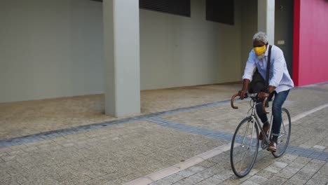 African-american-senior-man-wearing-face-mask-riding-bicycle-in-corporate-park