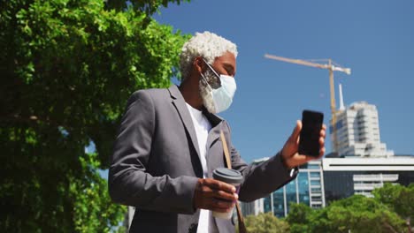 African-american-senior-man-wearing-face-mask-holding-smartphone-and-coffee-cup-hailing-taxi-on-the-