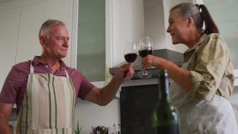 Caucasian-senior-couple-wearing-aprons-toasting-and-drinking-wine-in-the-kitchen-at-home