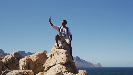 Afroamerikanischer-Mann-Beim-Wandern,-Der-Ein-Selfie-Macht-Und-Auf-Einem-Felsen-An-Der-Küste-Sitzt