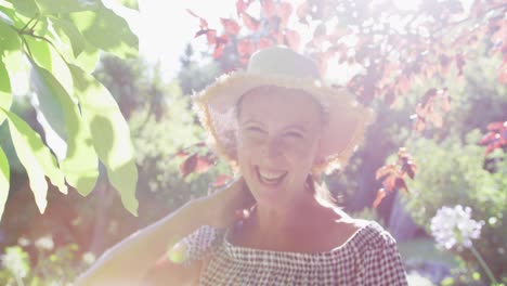 Retrato-De-Una-Mujer-Mayor-Caucásica-Feliz-Caminando-En-El-Jardín-Usando-Sombrero-Para-El-Sol-Y-Sonriendo-Bajo-El-Sol