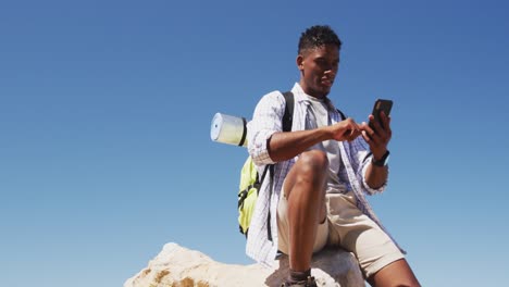 African-american-man-hiking-using-smartphone-sitting-on-rock-by-the-coast