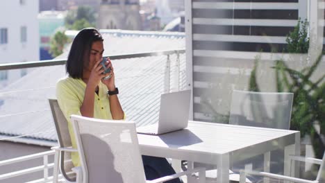 Mixed-race-gender-fluid-person-drinking-cup-of-coffee-and-using-laptop-on-roof-terrace