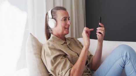 Kaukasische-ältere-Frau-Mit-Kopfhörern-Und-Smartphone,-Die-Zu-Hause-Auf-Der-Couch-Sitzt