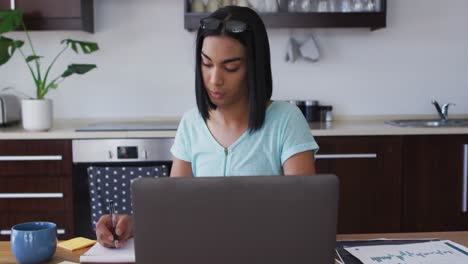 Mixed-race-gender-fluid-person-sitting-at-desk-taking-notes-working-from-home-using-a-laptop