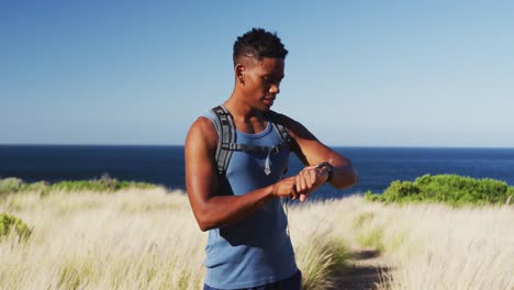African-american-man-cross-country-running-using-smartwatch-in-countryside-by-the-coast