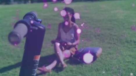 Animation-of-floating-purple-shapes-over-woman-sitting-on-grass-reading-book-in-park
