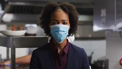 Portrait-of-african-american-female-manager-wearing-face-mask-in-restaurant-kitchen