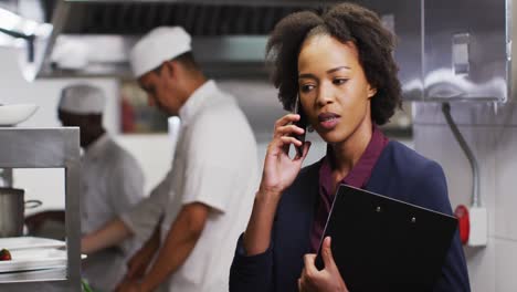 Gerente-Afroamericana-Hablando-Por-Teléfono-Inteligente-En-La-Cocina-Del-Restaurante