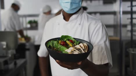 Portrait-of-african-american-male-chef-wearing-face-mask-presenting-dish