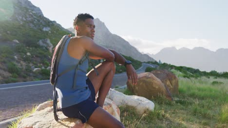 Afroamerikanischer-Mann-Beim-Cross-Country-Laufen,-Sitzend-Auf-Einem-Felsen-In-Der-Berglandschaft