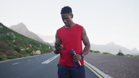 Hombre-Afroamericano-Sosteniendo-Una-Botella-De-Agua-Usando-Un-Teléfono-Inteligente-En-La-Carretera