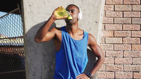African-american-man-wearing-headphones-drinking-from-water-bottle-taking-break-in-exercise-outdoors