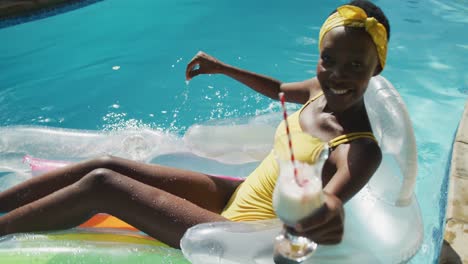 Attraktive-Afroamerikanische-Frau-Sitzt-Auf-Einem-Schlauchboot,-Hält-Ein-Getränk-In-Der-Hand-Und-Lächelt-Im-Schwimmbad