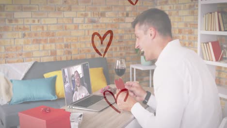 Heart-icons-over-caucasian-man-holding-a-heart-shaped-placard-while-having-a-video-call-on-laptop