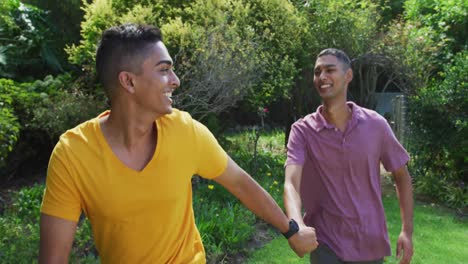 Smiling-mixed-race-gay-male-couple-holding-hands-walking-in-garden