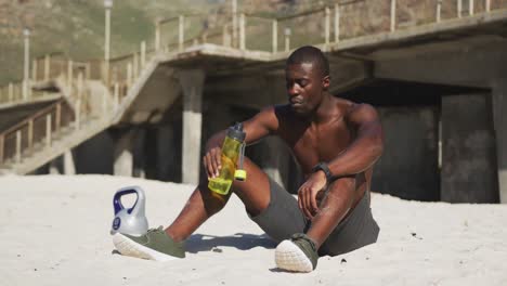 Tired-african-american-man-sitting-on-beach,-drinking-from-bottle,-taking-break-in-exercise-outdoors